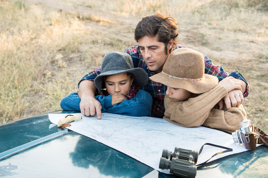 A father and two children leaning on a car watching a map in the forest