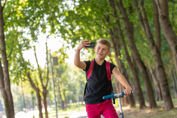 young happy preteen boy riding a scooter in warm sunny summer day in the park and taking selfie on smartpone while riding on scooter © Georgii
