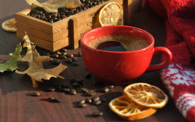 A red cup of coffee, a box of coffee, coffee beans, a warm knitted scarf and dry leaves are on the wooden table.