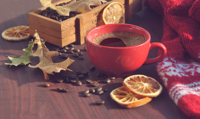 A red cup of coffee, a box of coffee, coffee beans, a warm knitted scarf and dry leaves are on the wooden table.