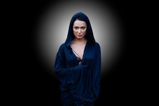 Young beautiful woman with a black hair and in the dark blue cloak with hood at the black background