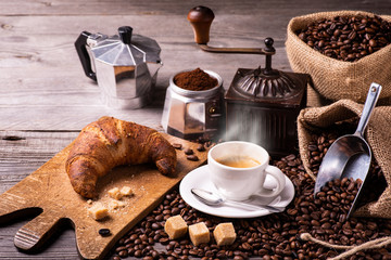 on the rustic wooden table a cup of hot coffee with croissant, a vintage coffee grinder, an Italian moka and coffee beans. High angle view