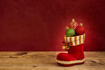 Ein roter Nikolaus Stiefel mit drei Christbaumkugeln auf einem Holzbrett vor einer roten Mauer