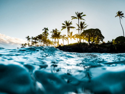 Beautiful Tropical Island Paradise Photo from Swimming In Clear Aqua Blue Ocean Water with Colorful Sky and Orange Clouds at Sunrise with Sun Rays Coming Through Bright Green Palm Trees in Maui Hawaii