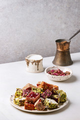 Variety of traditional turkish dessert Turkish Delight different taste and colors rose petals and pistachio nuts on ceramic plate with coffee jezve and milk jug on white marble table