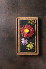 Beef tartare with quail egg in shell, cutting pickled cucumbers, capers, red onion, chives served on wooden black slate board with pepper mill on dark texture background. Flat lay, space