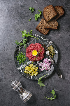 Beef tartare with quail egg in shell, cutting pickled cucumbers, capers, red onion, chives, bread, arugula salad served in marble plates with pepper mill on black texture background. Flat lay, space