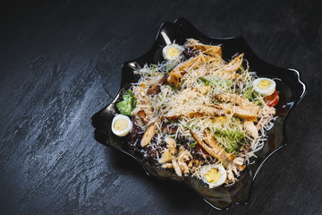 Delicious fresh caesar salad with chicken and eggs in a black plate on a black stone table background