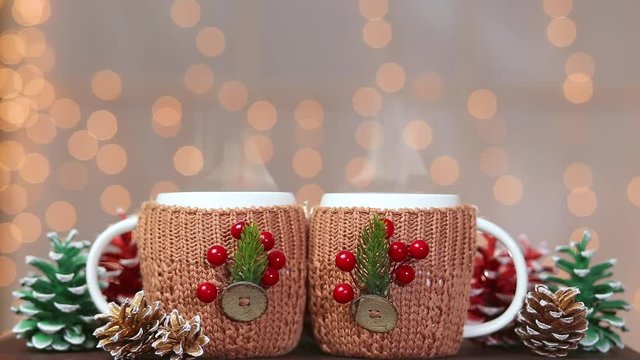 Two white mugs with hot drinks isolated on sparkling home background. Cups covered with cute knitted brown holders decorated with holiday Christmas and New year decorations. Real time full hd video.