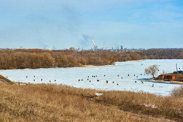 View to frozen river with fishermen catching fish on ice and factory with smoking chimneys on horizon. Environment pollution.