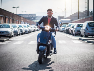 Young stylish man in suit and sunglasses driving scooter on empty road in sunny city.