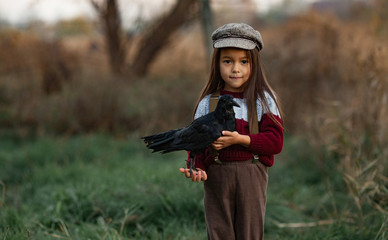 Child girl stands with raven in her hands on background of green grass.