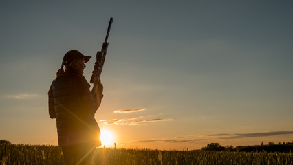Silhouette of a woman with a rifle. It stands in a picturesque place at sunset. Sports shooting and hunting concept - obrazy, fototapety, plakaty