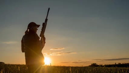 Deurstickers Silhouette of a woman with a rifle. It stands in a picturesque place at sunset. Sports shooting and hunting concept © StockMediaProduction