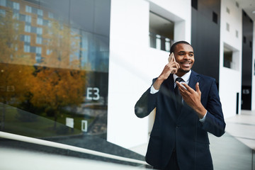 Happy young African businessman in suit talking by smartphone in urban environment