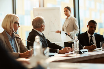 Mature delegate in formalwear paying attention to what young speaker by whiteboard saying