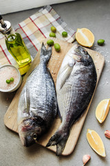 Fresh raw dorado or sea bream fish on a wooden kitchen board with lemon slices, spices, Himalayan pink salt, olive oil. Gray background. Mediterranean Kitchen.