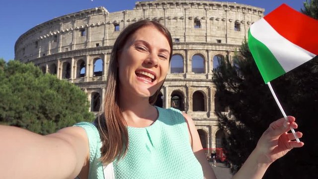 Woman doing selfie near famous attraction Colosseum in Rome, Italy. Teenage girl waving Italian flag in slow motion. Happy female tourist enjoying her european vacation. Student travel through Europe
