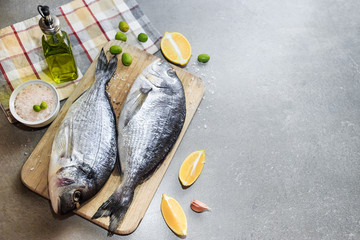 Fresh raw dorado or sea bream fish on a wooden kitchen board with lemon slices, spices, Himalayan pink salt, olive oil. Gray background. Mediterranean Kitchen. Top view. Place for text.