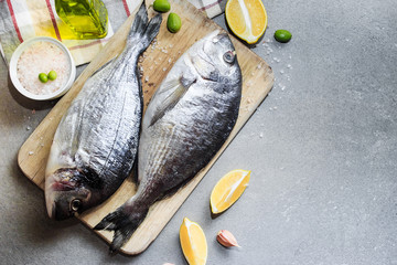 Fresh raw dorado or sea bream fish on a wooden kitchen board with lemon slices, spices, Himalayan pink salt, olive oil. Gray background. Mediterranean Kitchen. Top view. Place for text.