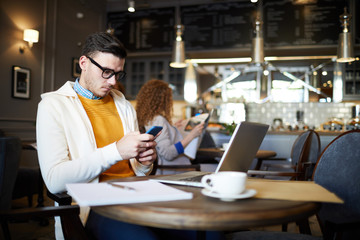 Casual young businessman scrolling in smartphone while sitting in cafe in front of laptop