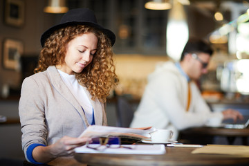 Fototapeta premium Young stylish wavy-haired businesswoman in hat reading the latest news in newspaper while sitting in cafe