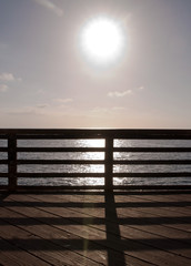 The sun brightly shining on the Pacific ocean seen through a wooden railing on a pier in southern California, USA