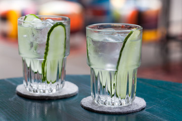 Gin tonic cocktail with ice and cucumber in a glass on a wooden table of a street cafe at a colorful blurry background