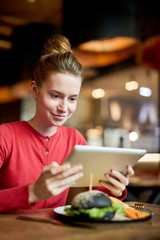 Pretty girl with touchpad sitting by served table in cafe and watching movie or video in the net