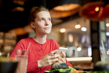 Pretty young pensive woman with smartphone scrolling or messaging while sitting in cafe