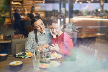 Girl with smartphone showing her friend curious promo while both sitting by table in cafe and...