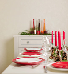 Merry Christmas and Happy New Year! Тable setting festive decor - white and red dishes, red candles in the candlesticks, juniper branches and fir cones.