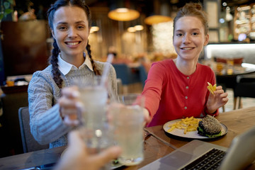 Cheerful girls in casualwear clinking with glasses of soda during lunch in fast food cafe