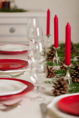 Merry Christmas and Happy New Year! Тable setting festive decor - white and red dishes, red candles in the candlesticks, juniper branches and fir cones.