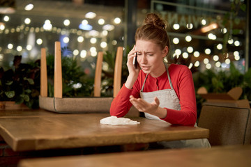 Upset waitress sitting by table and talking to someone by smartphone at the end of working day