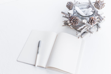 Open blank notebook, pen and handmade decoration candlestick with pine cones and branches in white tones.