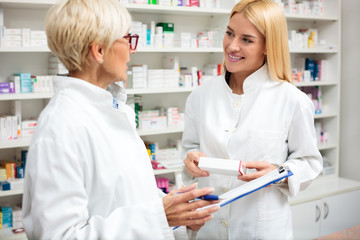 Obraz premium Happy young and mature blond haired female pharmacists standing in front of the shelves with medications, discussing. Writing on a clipboard and holding box of medications.