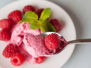 Closeup of a full spoon of raspberry ice cream and a tasty ripe berry above a blurred light background of a table with plate