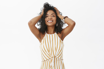 Girl feeling beautiful and feminine today. Charming happy and delighted stylisht tender african american female playing with curly hair and smiling satisfied at camera posing over gray wall