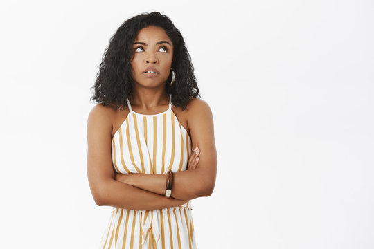 Arrogant and impatient young attractive african american woman in yellow striped overalls holding arms crossed on chest rolling eyes up from annoyance or irritation standing displeased over grey wall