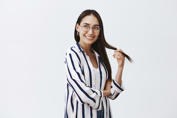 Studio shot of beautiful natural female student in glasses and blouse over t-shirt, playing with hair and gazing with dreamy and joyful expression left, smiling broadly over gray background