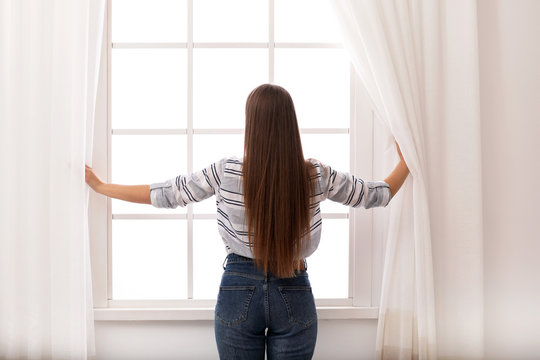 Young woman opening window curtains at home