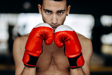 Sportsman with a naked torso keeps his hands in the red boxing gloves next to his face in the...