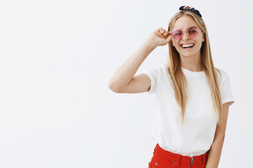 Waist-up shot of trendy cool and happy female celebrity in stylish sunglasses and headband touching rim of shades and smiling joyfully at camera having amazing trip on summer vacation over gray wall
