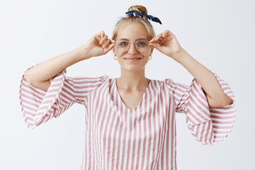 Waist-up shot of creative and stylish female model with fair combed hair wearing glasses and smiling joyfully attending optician picking eyewear and smiling broadly over gray background
