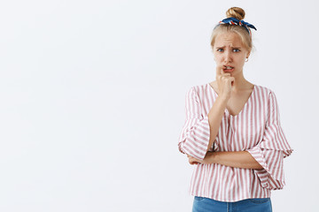 Studio shot of silly concerned and troubled insecure fair-haired woman with combed hair in striped blouse biting fingernail, frowning and grimacing feeling nervous and worried over gray background