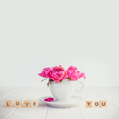 Close up light pink peony flowers bouquet in a decorative cup and saucer and message I love you in wooden blocks on white wooden background. Valentines day, good morning concept. Square. Copy space.