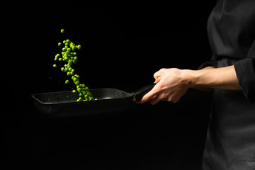 Professional cook. chef preparing dish with green peas in a pan. on a black background. menu, recipe book, healthy food, restaurant business