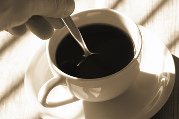Man stiring a cup of black coffee in sunlight on a wooden table with colour toning