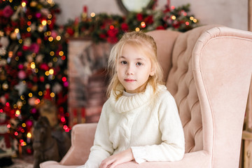 Christmas portrait of happy blonde child girl in white sweater siting on the floor near the Christmas tree. New Year Holidays